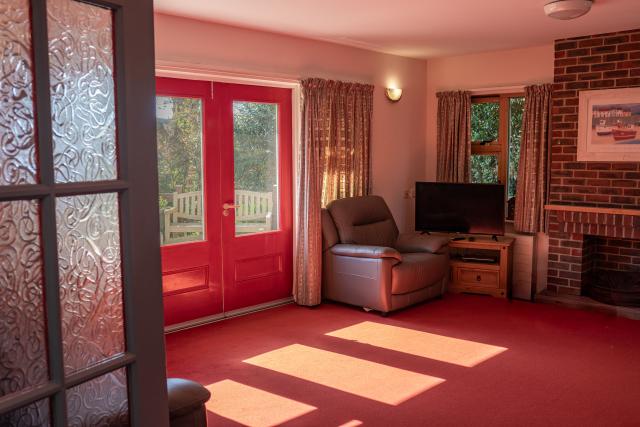 The living room at Barclay House, showing a red door and matching carpet opening into a wide open room, complete with a cosy chair, fireplace and television.
