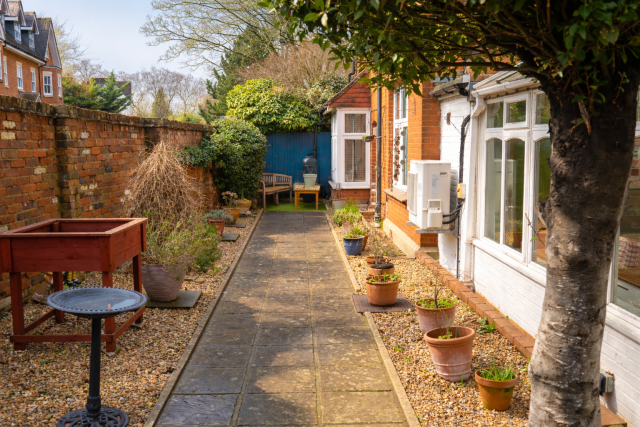 The back garden at Denecroft. Set next to the conservatory, there's a long central pathway flanked by pebbled borders, potted plants, and a bird fountain.