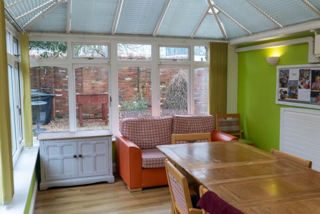 The dining area at Denecroft, set within a large conservatory and showing a long wooden table with a sofa at one end of the room.