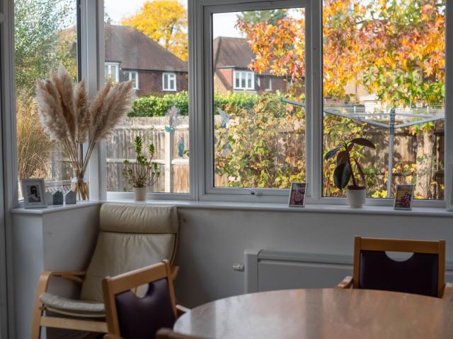 The dining area at Surbiton, showing a circular wooden table surrounded by chairs. The room is surrounded by bright glass windows, providing a view into the spacious garden outside.