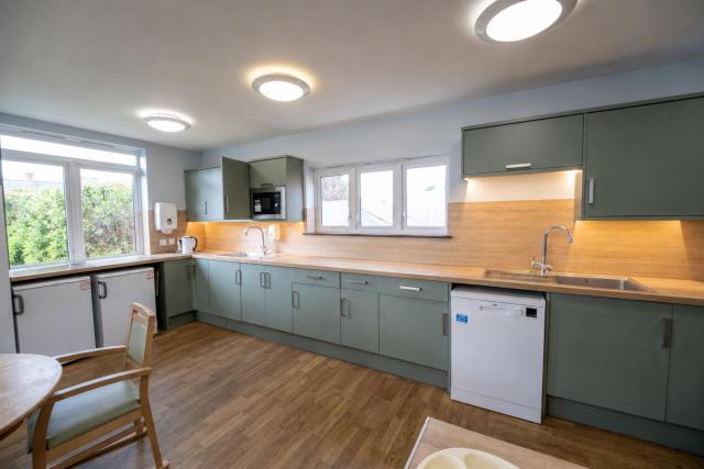 A kitchen at The Willows, showing a long kitchen surface with cupboards, a microwave, a dishwasher, and two sinks. There's a wide open space and a wooden table can be seen where people can enjoy their food. 